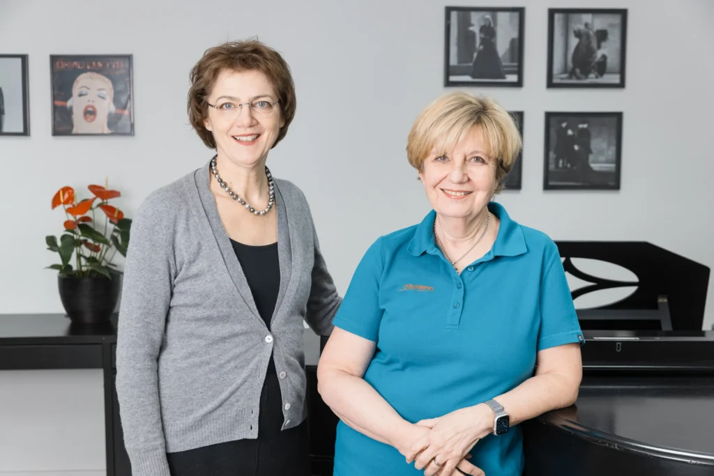 Dr. med. Salome Zwicky und Barbara Böhi stehen nebeneinander neben einem Klavier und lachen in die Kamera.