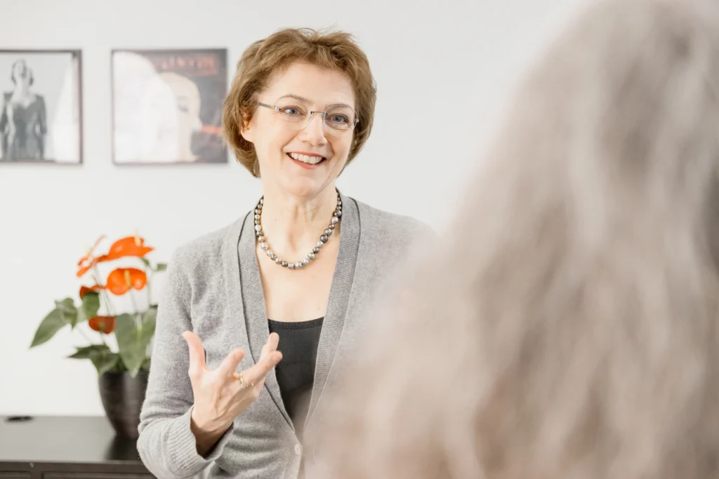 Eine Gesangspädagogin erklärt lächelnd etwas, während sie sich einer Patientin zuwendet.