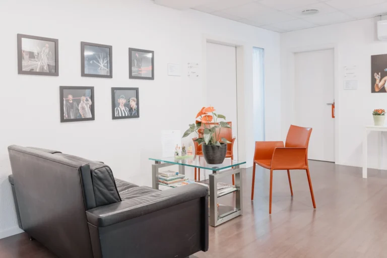 Wartebereich mit Sofa, rotem Stuhl und Pflanze; an der Wand hängen gerahmte Bilder von singenden Personen